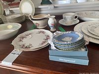 Wedgwood blue jasperware saucers stacked inside a Wedgwood box on the table alongside floral decorated small vase, Royal Albert serving dishes, Royal Ascot gravy boat, and several other plates and cups.