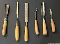 Photo showing six antique woodworking chisels with wooden handles and metal blades laid flat.