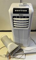 Front view of white Haier portable air conditioner with accessories laid out, showing main unit, exhaust hose and window vent panel.