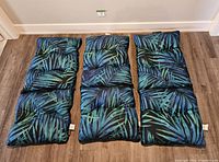 Three high back patio chair cushions laid flat in a row, showing one reversible side with large blue and green palm leaf pattern on black background.