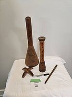 Four antique wooden sewing and textile tools arranged on a white cloth background including a tall textile spool, a bobbin, a slender quill, and a small X-shaped intersecting wooden piece, shown with a black marker for scale.