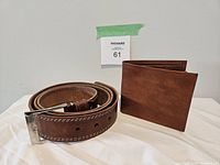 Photo of camel leather belt coiled with silver buckle and brown leather bifold wallet standing next to it.