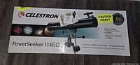Boxed Celestron PowerSeeker 114EQ 114mm reflector telescope showing the product image and brand logo on top, with caution heavy sticker and some minor box damage.