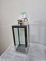 Chrome finished metal and glass lantern photographed on white surface against a white wall, showing front and side with door closed.