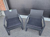 Pair of black patio chairs with metal frames and resin wicker seats and backs, each with removable black cushions, placed outdoors on pavement against dark backdrop.