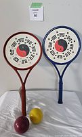 Front and partly side view of 2 rackets with yin-yang design and one yellow sand weighted ball, showing full racket face and handle details.
