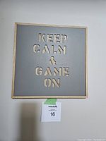 Square wall art with natural wood frame, grey laser cut panel, and phrase 'KEEP CALM & GAME ON'. Item is mounted on a white wall with a label below it.