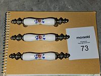 Three white ceramic drawer pulls with floral decoration on a yellow notebook with a lot card.