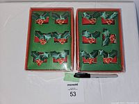 Front view of two unopened boxes of six holly napkin rings each, showing the green holly leaves with red berries inside green foam insert. Plastic wrapping intact with tape seals.