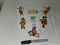 All six wooden Christmas figurines arranged on fabric background with black marker for scale. Four figurine ornaments and two wooden accessory items visible with some straw details and colorful felt clothing.