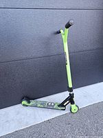 Full side view of green and black Vokul VK3 Pro youth stunt scooter standing against a dark wall on pavement, showing overall condition and design.
