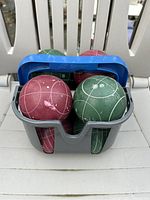 Eight bocce balls (four red, four green) with white markings in a gray plastic carrying case with blue handle positioned on a plastic chair seat.