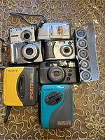 Top down view of cameras and accessories laid out on a patterned golden cloth background. Includes digital and film cameras, two Sony Walkman Sports cassette players, and seven rolls of film in a holder.