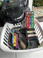Black portable CD player resting on the edge of a white basket containing multiple CDs and VHS tapes.