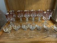 Photo showing 8 amethyst goblet-style glasses and 7 small clear glass cups with handles on a wooden surface.