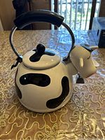 Front and side view of the cow design tea kettle showing black spots and a spout shaped like a cow's head.