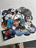 Photo showing a collection of loose Xbox and PlayStation video game discs scattered on a white surface. Some discs have recognizable cover art and titles visible like The Matrix, Resident Evil, and Project Gotham Racing.