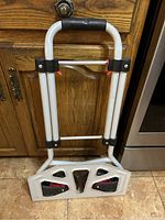 Folding dolly hand truck folded against a wooden kitchen cabinet door and tiled floor.
