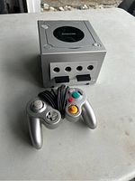 Silver Nintendo GameCube console with one wired controller placed in front, showing front ports and controller cables.