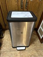 Garbage bin front view showing stainless steel body, black top with white marble-like panel, and foot pedal.