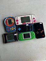 Six handheld electronic games arranged on a surface, showing front screens and control buttons distinct by color and branding.