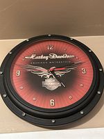 Harley Davidson branded wall clock with red dial and black rim, showing clock hands and Harley Davidson logo.