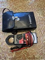 Hioki clamp meter tester with red and black leads coiled, resting on a patterned surface, alongside a black carrying case with clasp and white Hioki logo.