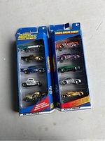 Two unopened blister card packs with five Hot Wheels die-cast cars each, set on a concrete surface with visible details of car paint and decals.