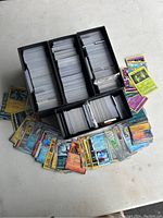 Image showing multiple black card storage boxes filled with sleeved Pokemon trading cards, with many loose cards spread around the boxes on the ground.