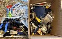 Top view of box showing variety of office supplies and small hand tools including scissors, wire cutters, stapler, putty knives, and assorted hardware pieces.
