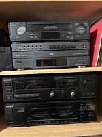 Front view of a shelving unit holding a stack of four audio/video equipment devices: one Sharp VHS VCR and three Sony audio components including a CD player, an AV control center, and a cassette tape deck. The shelf also shows part of some vinyl records or large flat items on the right side.
