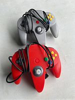 Photo of two Nintendo 64 wired controllers, one in red and the other in grey, showing front-facing analog sticks and buttons with wires coiled loosely around them.
