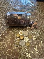 Glass jar filled with many old coins, some spilled out next to it on an embroidered fabric surface.