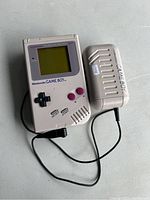 Front view of original Nintendo Game Boy with AC adapter plugged in, showing screen, controls, and overall console condition.