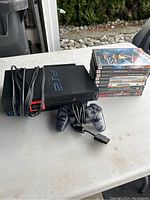 Photo showing PlayStation 2 console, wired controller and cables, with stack of 8 game cases and discs to the side on a light colored table outdoors.