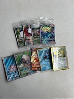 The image shows four sealed Pokemon card packs in clear plastic packaging along with eight loose Pokemon cards spread out below them on a light surface.