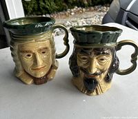 Two Royal Doulton large mugs sculpted as human faces with vivid expressions and detailed features, placed outdoors on a table.