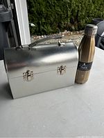 Silver tin lunch box with handle and latches standing next to a stainless steel insulated bottle on a light surface outside.