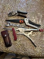 Photo showing six assorted hand tools on a quilted patterned surface: wire cutter, adjustable wrench, locking pliers, pocket knife, staple gun, and leather sheath.