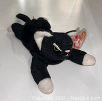 Photo showing a black cat Beanie Baby plush toy lying down with green eyes, white paws, and a red TY heart tag.