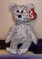 White plush teddy bear decorated with silver stars, black eyes and nose, Ty tag from year 2000 attached to the ear, displayed against beige wall.