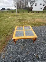 Image of a square wooden coffee table with a glass pane top placed outdoors on a gravel surface with a lawn and house in the background.