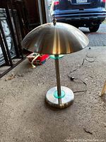 Brushed metal desk lamp with dome shade and green glass accents on base and stem, shown with power cord plugged in.