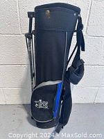 Photo du sac de golf noir avec support pliant et poche latérale bleue et logo 'Sticks for Kids' visible.