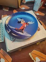 Photo of a decorative plate featuring Mickey Mouse as the Sorcerer's Apprentice on a blue background, placed on a cardboard box.