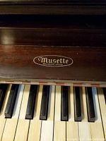 Close-up photo of the Musette brand emblem on the piano above the keyboard showing wooden finish and keys.