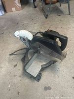 Portable table saw on concrete floor showing black and gray body, circular saw blade, handle, and attached white dust collection hose.