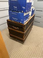 Large vintage wooden trunk with metal reinforcements and latch, visible caster wheels at bottom, placed in a small room with boxes on top.