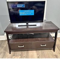 Wooden TV stand holding a flat screen TV, showing the front with open shelf and two pull-out drawers with metallic handles.