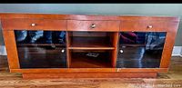 Wooden TV stand with three drawers with silver knobs and two glass doors around a central open shelving space.